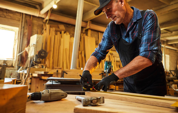 Portrait d'un menuisier senior travaillant avec un bois, marquant la planche avec un crayon et prenant des mesures pour couper un morceau de bois pour faire un meuble dans l'atelier de menuiserie - Photo, image