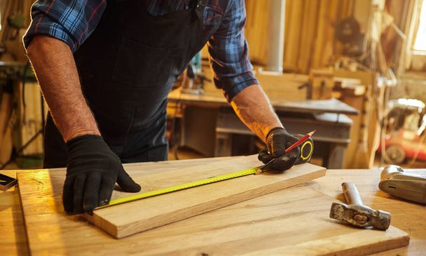 Carpenter working with a wood, marking plank with a pencil and taking measurements to cut a piece of wood to make a piece of furniture in a carpentry workshop, close-up view - Foto, afbeelding