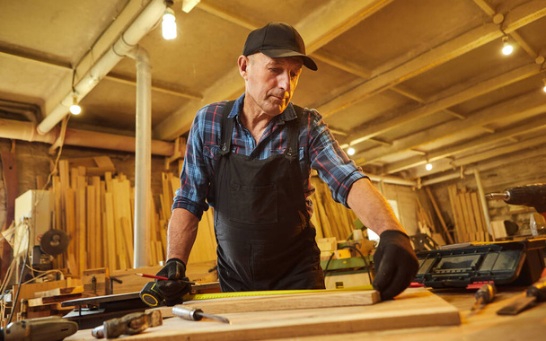 Προσωπογραφία ενός παλαιού ξυλουργού που εργάζεται με ένα ξύλο, σήμανση σανίδα με ένα μολύβι και τη λήψη μετρήσεων για να κόψει ένα κομμάτι ξύλου για να κάνει ένα κομμάτι των επίπλων στο εργαστήριο ξυλουργικής - Φωτογραφία, εικόνα