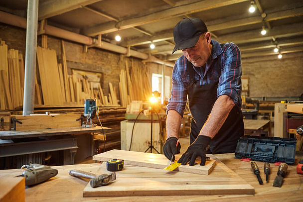 Portret van een senior timmerman die met een hout werkt, planken markeert met een potlood en afmetingen neemt om een stuk hout te snijden om een meubelstuk te maken in de timmerwerkplaats - Foto, afbeelding