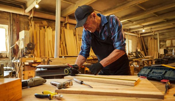 Портрет старшего плотника, работающего с деревом, маркирующего доску карандашом и делающего замеры, чтобы обрезать кусок дерева, чтобы сделать мебель в столярной мастерской - Фото, изображение
