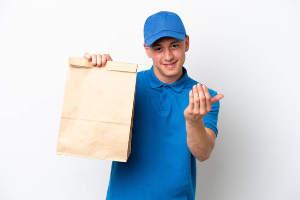 Young Brazilian man taking a bag of takeaway food isolated on white background inviting to come with hand. Happy that you came - Zdjęcie, obraz