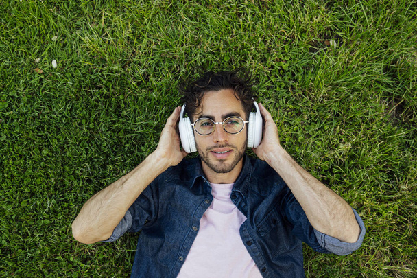 Εξωτερικό πορτραίτο ενός όμορφου νεαρού hipster άντρα ξαπλωμένος στον κήπο με ακουστικά. - Φωτογραφία, εικόνα