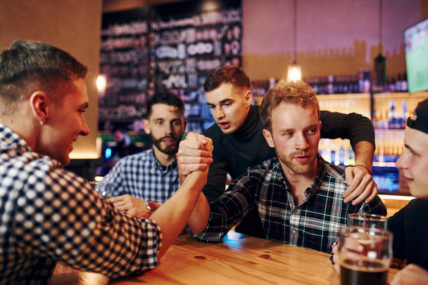 Friends plays arm wrestling. Group of people together indoors in the pub have fun at weekend time. - Foto, immagini