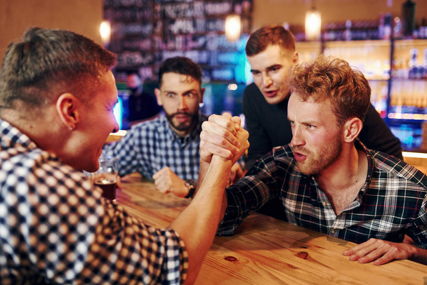 Friends plays arm wrestling. Group of people together indoors in the pub have fun at weekend time. - Foto, afbeelding