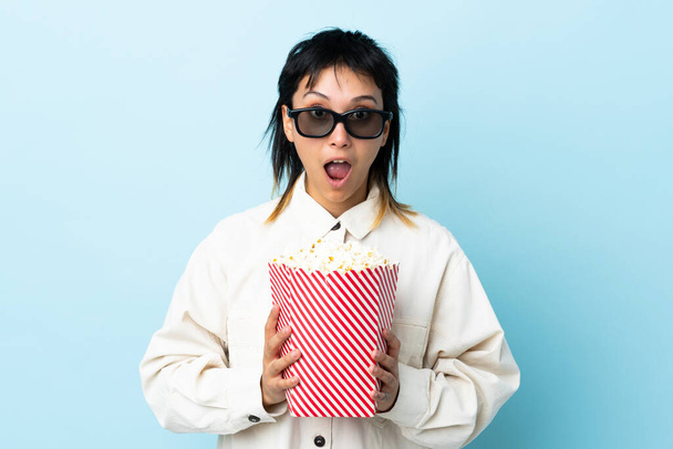 Young Uruguayan woman over isolated blue background surprised with 3d glasses and holding a big bucket of popcorns - Photo, Image