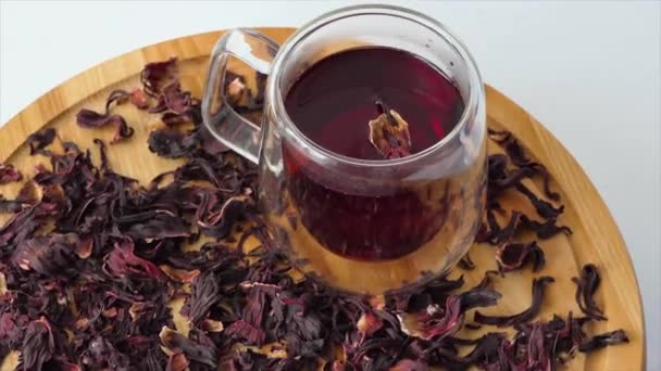 Červený čaj ve skleněném šálku na bílém pozadí. Červený ibiškový čaj se nalévá do průhledné sklenice s dvojitými stěnami. Šálek čaje rotující na dřevěné desce. - Záběry, video