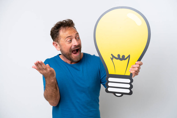Middle age caucasian man isolated on white background holding a bulb icon with surprised expression - 写真・画像