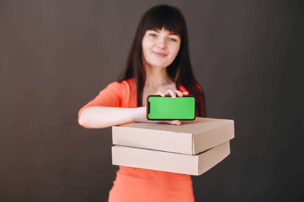 In the foreground are flat boxes and above them a phone with a green screen in the hands of a cheerful girl - Foto, Imagen
