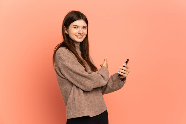 Young Ukrainian girl isolated on pink background using mobile phone and pointing back - Foto, afbeelding