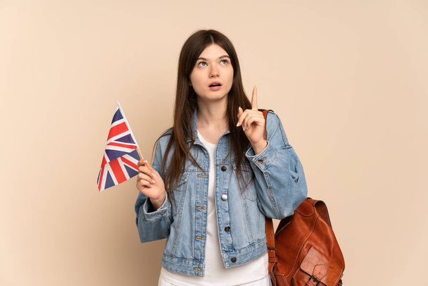 Joven ucraniana sosteniendo una bandera del Reino Unido aislada sobre fondo beige pensando en una idea apuntando con el dedo hacia arriba - Foto, Imagen