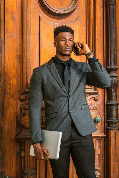 Hombre de negocios afroamericano con perilla trabajando en Nueva York. El uso de la chaqueta de moda, corbata negro, la celebración de ordenador portátil, chico joven, de pie por la puerta de estilo vintage, llamando por teléfono - Foto, imagen