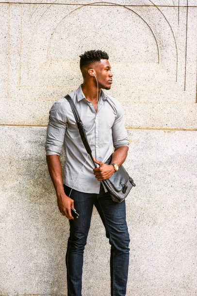 Homem Africano Americano viajando, trabalhando em Nova York, vestindo camisa, jeans, relógio de pulso, carregando bolsa de couro de ombro, na rua, ouvindo música com fone de ouvido e telefone celular. efeito filtrado - Foto, Imagem