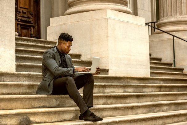 African American Businessman traveling, working in New York, wearing fashionable jacket, necktie, sitting on stairs outside office building, reading, working on laptop computer. Color filtered effect - Foto, afbeelding