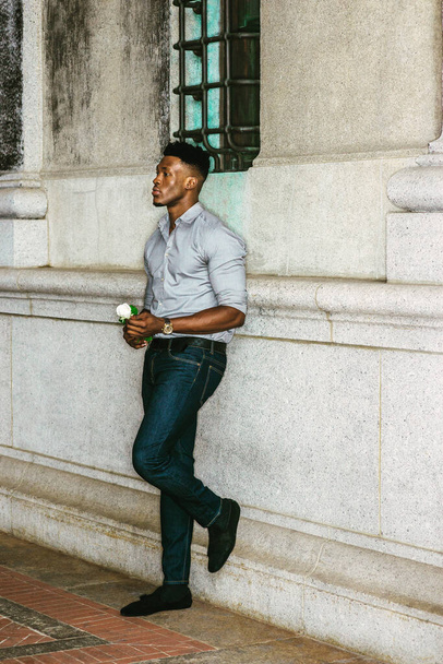 Afrikai amerikai férfi, aki szerelmet keres New Yorkban, szürke inget, kék farmert, cipőt, karórát visel, falon áll az utcán, fehér rózsát tart a kezében, gondolkodik. Hiányzol. szűrt hatás - Fotó, kép