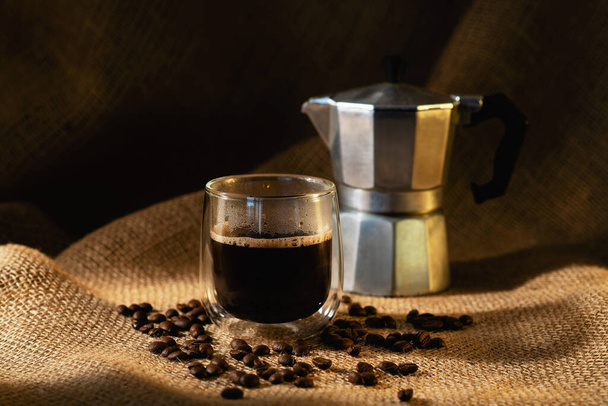 A transparent glass cup of coffee and a geyser coffee maker on a dark burlap background with coffee beans. Horizontal photo. - Fotografie, Obrázek