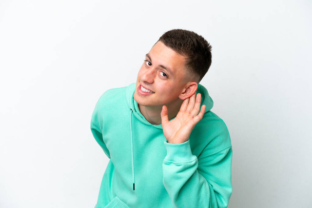 Joven brasileño aislado sobre fondo blanco escuchando algo poniendo la mano en la oreja - Foto, Imagen