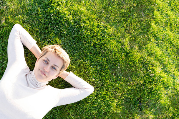 Femme aux cheveux courts en haut blanc reposant sur l'herbe verte de prairie par une journée chaude ensoleillée de printemps dans le parc - Photo, image