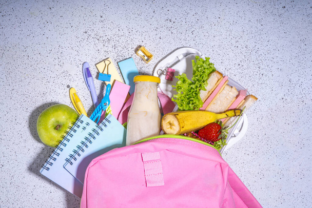Zdrowe pudełko śniadaniowe szkoły: kanapki, warzywa, owoce, orzechy i jogurt z zaopatrzeniem dla dzieci w szkole, akcesoria i plecak na czarnym tle płaskiej przestrzeni kopii. Powrót do koncepcji szkoły - Zdjęcie, obraz