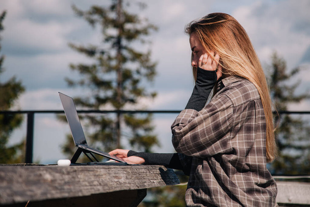 A young, slender girl in a plaid shirt works at a laptop in nature against the backdrop of mountains - Fotografie, Obrázek