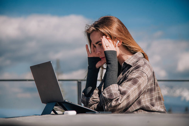 A young, slender girl in a plaid shirt works at a laptop in nature against the backdrop of mountains - Foto, afbeelding