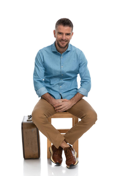 bearded man in his forties with luggage going to vacation, sitting on wooden chair, smiling and touching hands in front of white background in studio - Foto, afbeelding