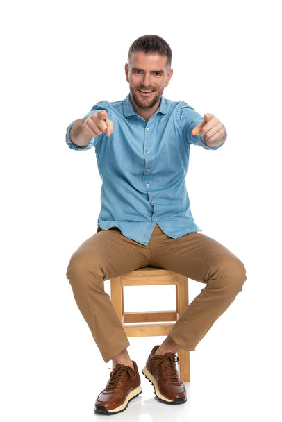 full body picture of attractive middle aged man in denim shirt with chino pants and sneakers smiling and pointing fingers whil sitting on wooden chair on white background - Foto, Imagen