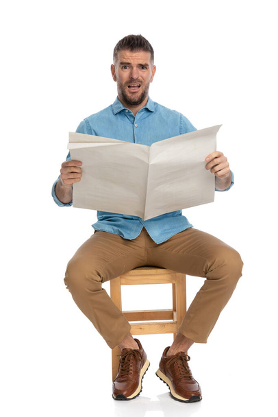 full body picture of bearded man with sneakers reading newspaper and grimacing while sitting on wooden chair on white background in studio - Foto, Imagem