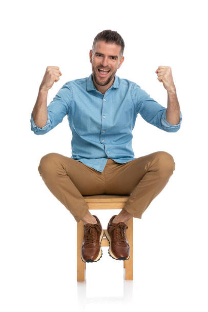 excited casual guy in denim shirt with beard holding fists up and cheering while sitting on white background, laughing and posing in studio - Foto, afbeelding