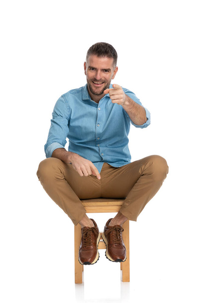 unshaved seated man in denim shirt, with chino pants and sneakers sitting on chair, pointing finger and smiling on white background - Foto, Imagem