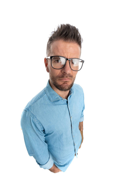 casual guy in his 40s with glasses holding hands in pockets and grimacing on white background in studio - Photo, Image