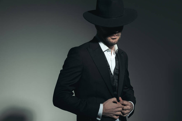 portrait of mysterious best man in tuxedo with hat covering face with hat while buttoning tux jacket on grey background - Photo, image