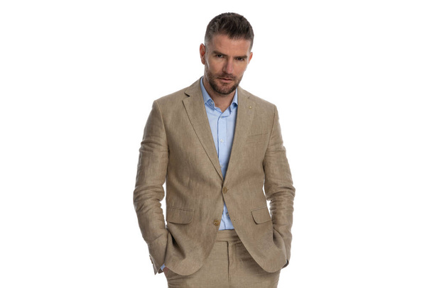 portrait of serious young businessman with undone shirt and beige suit holding hands in pockets in front of white background in studio - Fotoğraf, Görsel