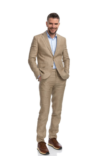full body picture of happy businessman in beige suit holding hands in pockets and smiling in front of white background in studio - Photo, Image