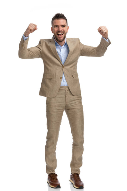 Ganzkörperbild eines glücklichen Mannes in seinen Vierzigern im beigen Anzug mit aufgelöstem Hemd, der die Arme in die Luft reckt und den Sieg auf weißem Hintergrund feiert - Foto, Bild