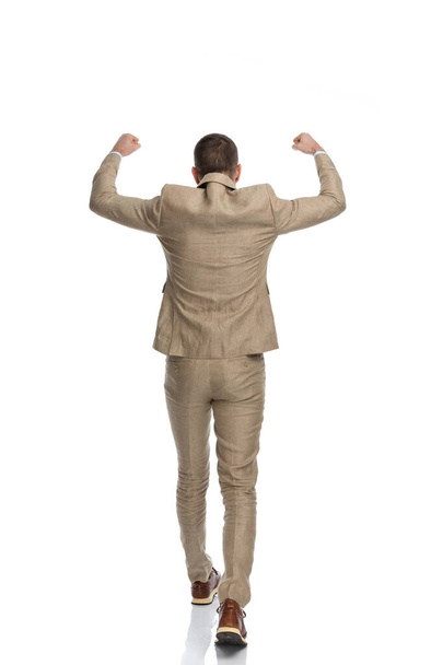 back view of businessman in beige suit holding arms in the air and walking while celebrating victory on white background in studio - Foto, afbeelding