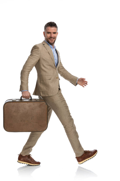 happy bearded man in his 40s with luggage quitting job and going to vacation, walking and smiling in front of white background - Photo, image