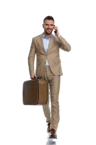 happy man in suit wearing undone shirt, holding luggage, talking on the phone, smiling and walking in front of white background in studio - Photo, Image