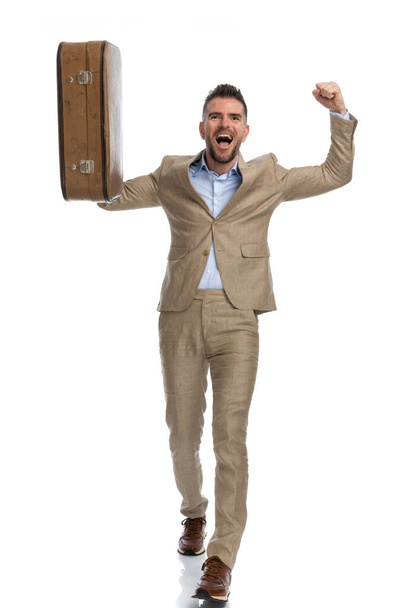 full body picture of man in his 40s with arms above head being enthusiastic for his long waited holiday, laughing and holding luggage on white background in studio - Foto, immagini