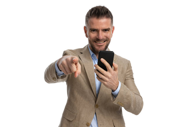 smiling young businessman in beige suit holding telephone and showing with pointed finger on white background - Photo, Image