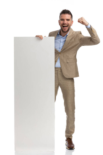 40 'lı yaşlarında coşkulu bir adam boş tahtayı tutuyor ve sunuyor ve havada yumruğuyla kutluyor, beyaz arka planda gülüyor. - Fotoğraf, Görsel