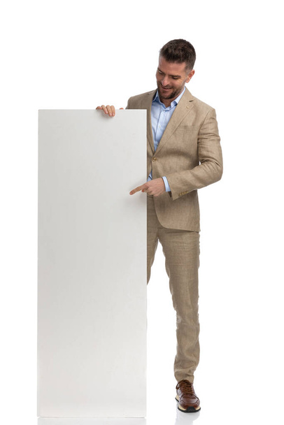 feliz hombre de mediana edad en traje beige con camisa deshecha apuntando con el dedo y mostrando tablero vacío sobre fondo blanco en el estudio - Foto, imagen