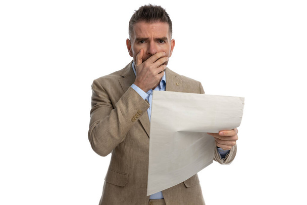 portrait of shocked man in beige suit reading newspaper and covering mouth with hand on white background - Фото, изображение