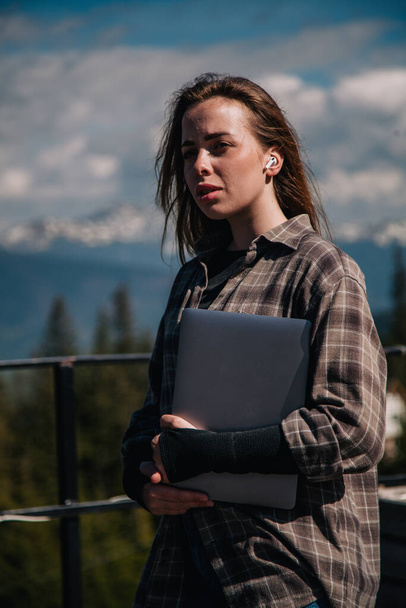 A young, slender girl in a plaid shirt with a laptop in her hands stands in nature against the backdrop of mountains. - Photo, image