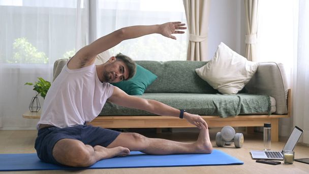Young man in sports clothes is warming up, stretching while sitting on mat at home - Φωτογραφία, εικόνα
