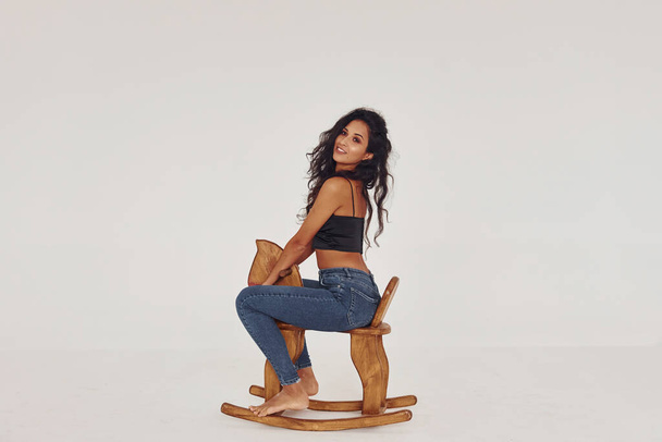 Sits on kid's wooden horse toy. Young woman in jeans and bra indoors. White background. - Zdjęcie, obraz