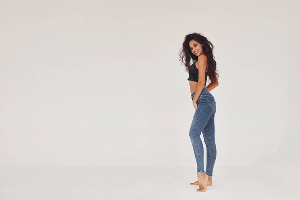 Curly hair. Young woman in jeans and bra indoors. White background. - Photo, image
