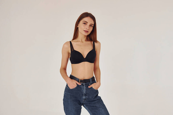 Young woman in jeans and bra indoors. White background. - Fotoğraf, Görsel