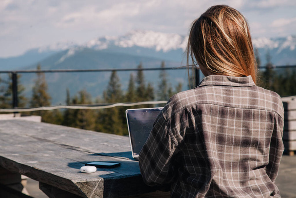 A young, beautiful girl in a plaid shirt works on a laptop at a wooden old table in nature against the backdrop of mountains, next to a smartphone and headphones. View from the back. - Zdjęcie, obraz