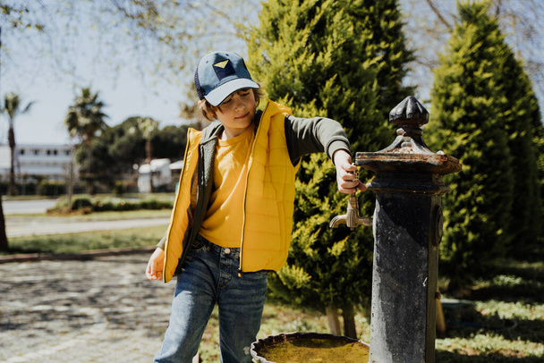 Kid schoolboy in casual clothing washing his hands in the street old fashioned drinking water fountain. Boy in blue jeans and yellow vest playing with water from drinking sprinkler in city park - Foto, Imagen